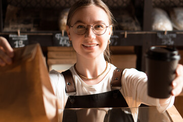 Young smiling woman salesperson in bakery holding paper bag and coffee cup to go in cafe