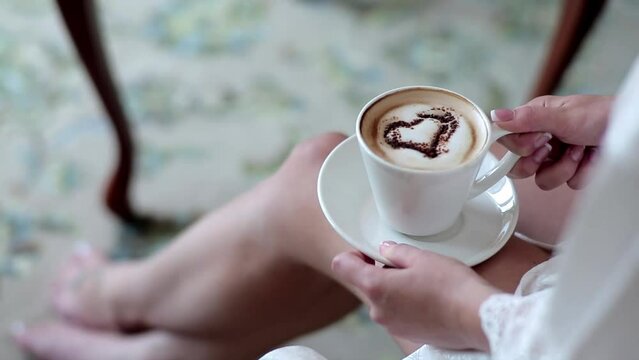 A cup of creamy cappuccino coffee with a foam in the form of a heart in a woman's hand. Bride drinking coffee