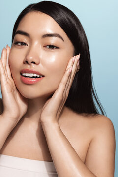 Beautiful Girl With Bare Shoulders Applying Cream On Her Face And Smiling Against Light-blue Background. Smiling Asian Woman With Glowing Skin Applying Facial Skincare Cream