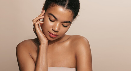 Beautiful african american woman with glowing, nourished head and shoulders, touches her moisturized, perfect facial skin after skincare product, brown background