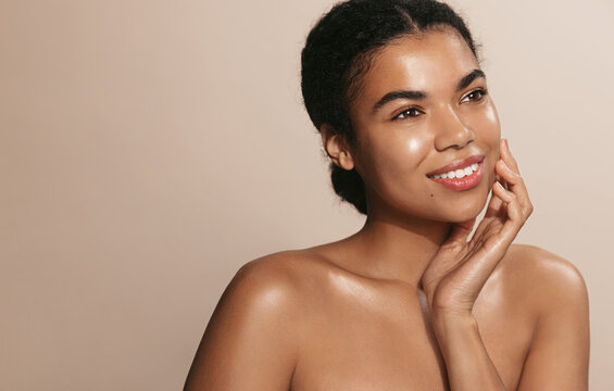 Beauty female model with nourished, glowing skin, smiling, gently touching her moisturized face, brown background