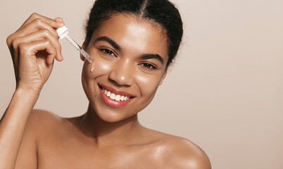 Smiling woman with perfect skin, applies tea trea, collagen face lifting serum, holds dropper, stands over brown background