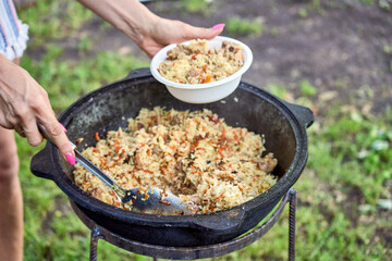 A woman spoons hot pilaf into a plate from a cauldron. Cooking pilaf in a cauldron on fire in the...