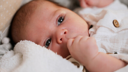 Active newborn baby lies in crib and moves and looks around and explores world. Calm boy or girl opens his mouth and pulls his fist into mouth. Child wants to eat. Paternity. Happiness of motherhood