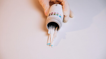 Close-up of hand in disposable medical glove pour out test strips from plastic jar on white background. Strips for urine diagnosis. Test for glucose and acetone. Strips for self-diagnosis. Laboratory