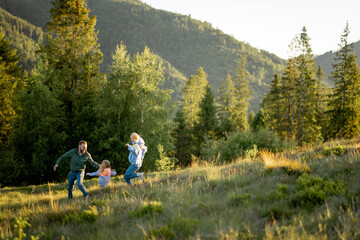 Young family with little girl walk together on green meadow while traveling in the mountains during sunset. Happy family spending summer vacation on nature