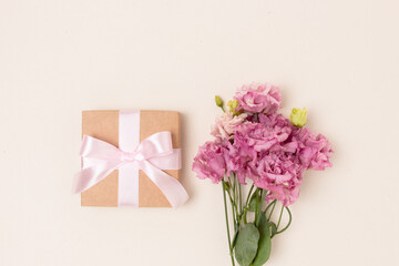 Gift box with tied bow and bouquet of eustoma flowers on a beige background. Springtime festive concept with copyspace.