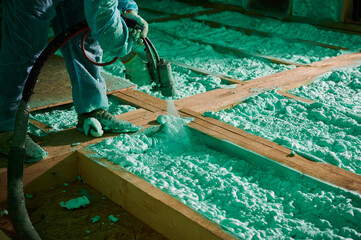 Male builder insulating wooden frame house. Cropped view of man worker spraying polyurethane foam on floor inside of future cottage, using plural component gun. Construction and insulation concept. - obrazy, fototapety, plakaty