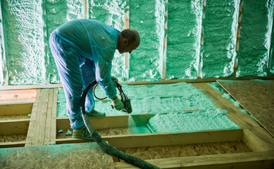 Male builder insulating wooden frame house. Man worker spraying polyurethane foam inside of future cottage, using plural component gun. Construction and insulation concept. - obrazy, fototapety, plakaty