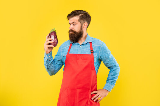 Serious Man In Red Apron Looking At Eggplant Yellow Background, Grocer