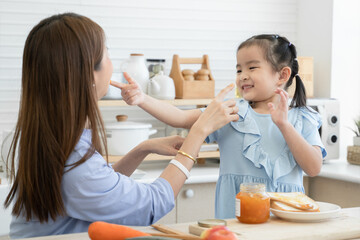 Obraz na płótnie Canvas Happy Asian family, cute little daughter and young mother enjoy teasing putting jam on each other face while preparing breakfast together with fruits and vegetables in kitchen at home