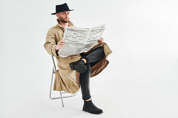 Young handsome man in a trench coat and hat reading newspaper sitting on chair over white background - obrazy, fototapety, plakaty