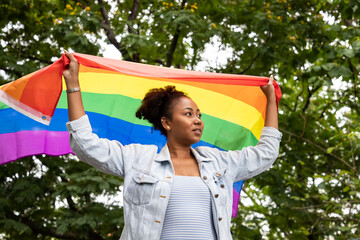 Portrait of young lesbian woman waving pride rainbow flag in their backs supporting LGBTQ pride in...