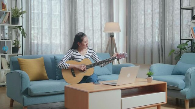Asian Woman Learning Pulling Strings Of Guitar By Laptop At Home
