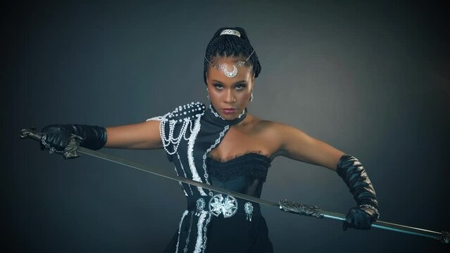 portrait african fantasy woman warrior queen holding sword in hands draws from scabbard. Beauty sexy face halloween makeup Carnival black dress white pearls. girl princess tiara moon, gray studio