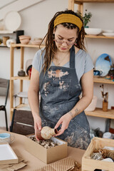 Young female in workwear taking handmade clay vase or jug out of box while standing by table with...
