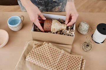 Above angle of hands of craftswoman putting handmade jug or vase into box while packing one of...