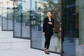 A full length photo of a lady in a blazer who is leaning against the glass wall in the financial district. A businesswoman with her arm on her chest in the contemporary urban space.