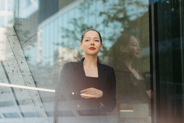 A close portrait of a lady in a blazer who is leaning against the glass wall in the financial district. A businesswoman with her arm on her chest in the contemporary urban space.