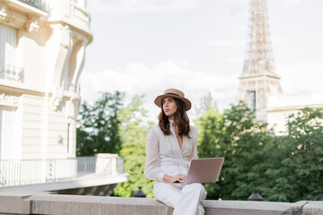 Trendy stylish freelancer in sun hat using laptop on street with Eiffel tower at background in Paris.