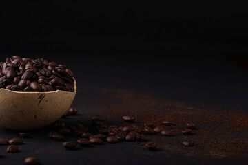 A bowl is full of coffee beans on a black surface with scattered coffee beans leaving space to...