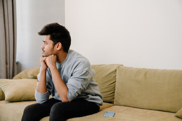 Young thoughtful indian man sitting on sofa and thinking