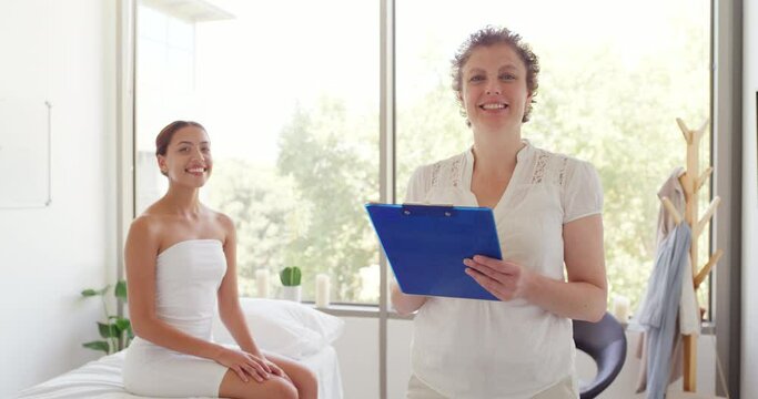 Portrait of a female doctor or masseuse taking notes on a clipboard while consulting with a patient in a clinic. A mature physiotherapist writing the history of her client sitting behind her