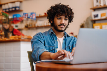 Young calm handsome indian man working with laptop at cafe