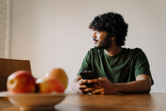 Young Indian Handsome Curly Man Sitting By Table With Phone