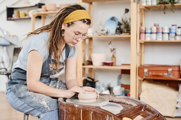 Young creative woman in casualwear and apron bending over pottery wheel and creating new...