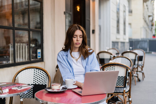Stylish freelancer using laptop near coffee and dessert on cafe terrace in Paris.