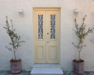 A house front with a pale white door and a wall decorated with potted olive plants. Travel to Tinos...