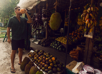 Male model, traveler drinking coconut against the backdrop of an exotic fruit shop, holiday landscape.