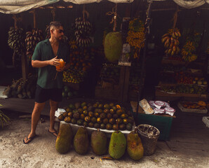 Male model, traveler drinking coconut against the backdrop of an exotic fruit shop, holiday landscape.