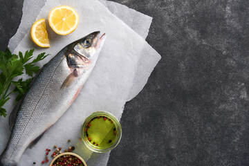 Sea Bass raw. Fresh sea fish bass with salt, pepper, parsley, olive oil and lemon on parchment paper on a dark concrete rustic background. Food cooking background. Top view, copy space.
