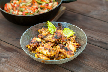 A frying pan with stewed vegetables on a wooden table next to a dish with ruddy grilled wings.