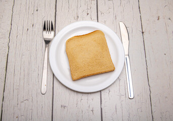 A single slice of toast on a plain white plate and cutlery with signs of use on a rustic wooden...