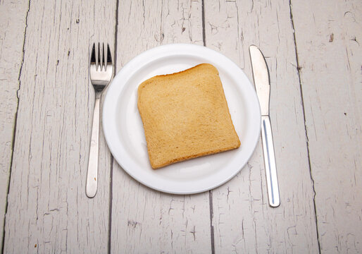 A single slice of toast on a plain white plate and cutlery with signs of use on a rustic wooden surface
