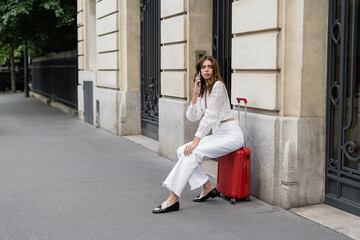 Young woman talking on smartphone while sitting on suitcase on urban street in Paris.