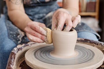 Fototapeta na wymiar Hand of female potter holding sponge close to rotating clay item on pottery wheel while creating new earthenware in workshop