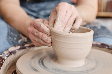 Hands of female owner of small earthenware shop creating new pot, bowl or other clay item while...