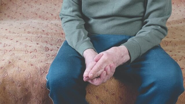 Medium shot of an old man rubbing his wrinkled hands while sitting on the couch. The man rubs his hands, stroking the bone on his arm. The hands of a pensioner. Selective focus, shallow depth of field
