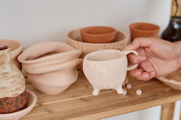 Hand of young female potter taking handmade clay cup from wooden shelf or display with assortment of earthenware for sale