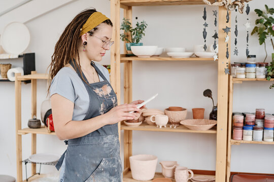 Side view of young creative female owner of earthenware shop using tablet while looking through new online orders of clients