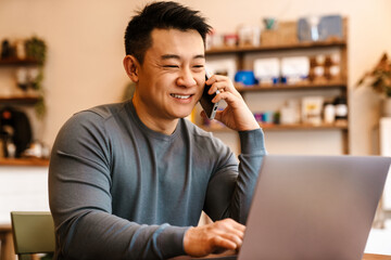 Happy asian man talking on cellphone and using laptop in cafe indoors