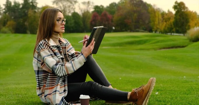 Beautiful young university or college student girl in glasses is sitting in the park, university campus, drawing on her tablet computer with electronic pen or pencil, studying online with her gadget