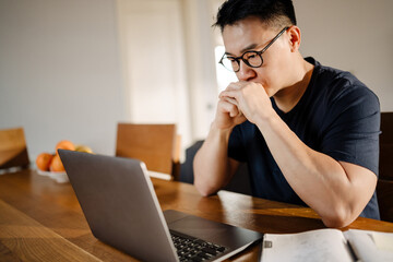 Serious adult asian man thinking while working on laptop at home