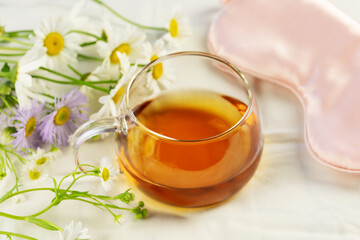 Morning, herbal tea. Concept of relaxing, self-care and good sleeping. Glass cup of tea, sleeping mask and wild flowers on a white bed linen. Summer, good morning. Soft focus style image