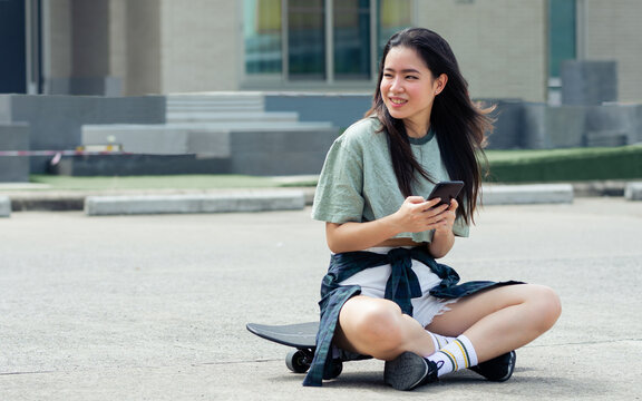 Portrait beautiful sportive Asian female skater wearing hipster clothes, smiling with happiness, sitting on skateboard outdoor, chatting on mobile phone with copy space. Activity and Adventure Concept