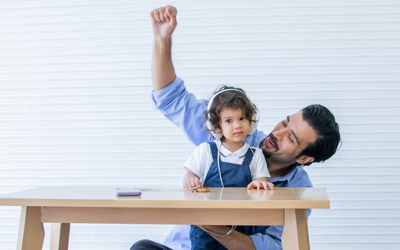 Caucasian Handsome Beard Father Or Dad Wearing Working Shirt With Necktie, Taking Headphone To His Little Adorable Curly Hair Little Daughter For Music At Home. Education, Family, Kid Concept.